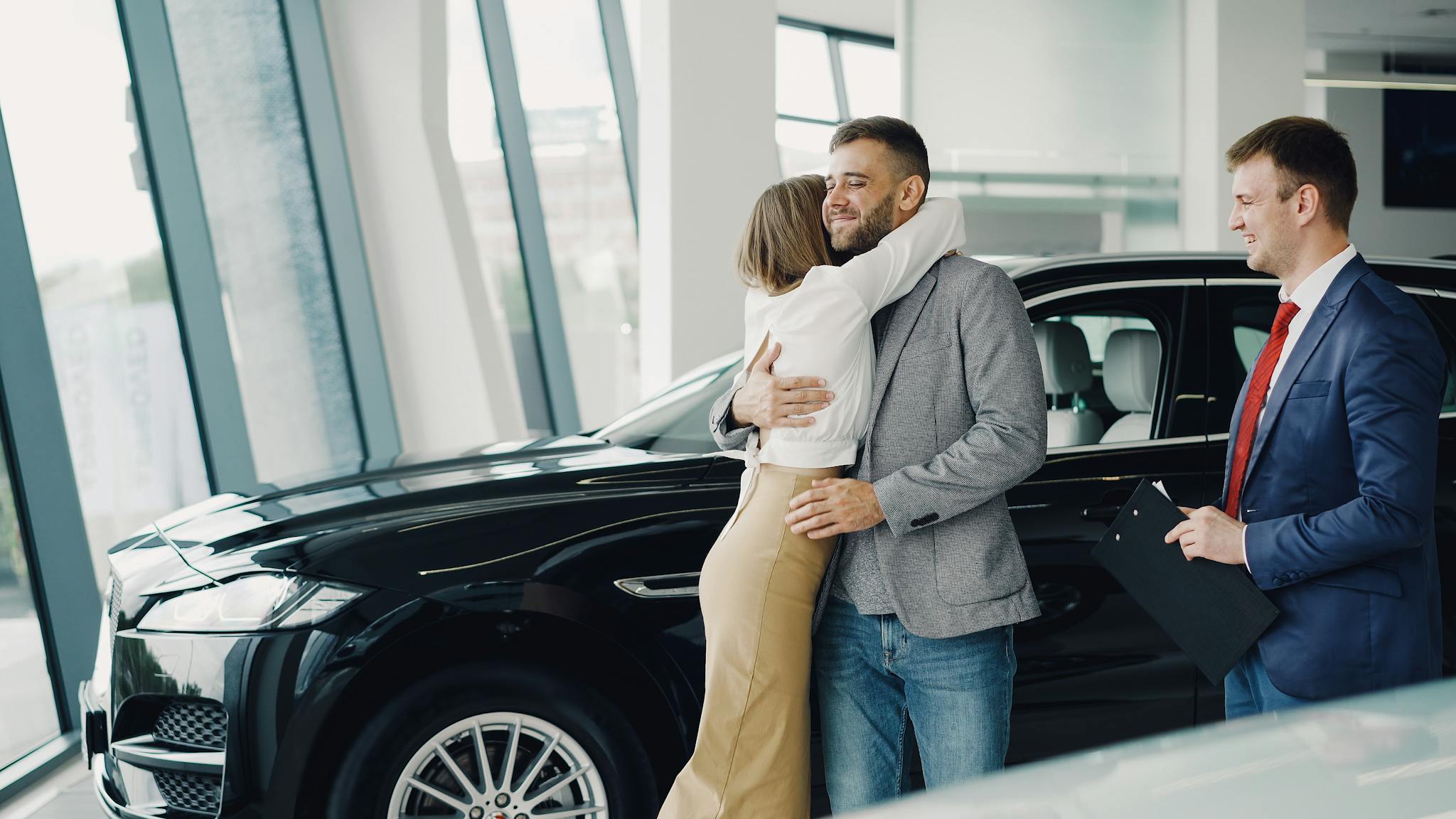 Couple hugging with car salesman after purchasing a new car at a dealership.